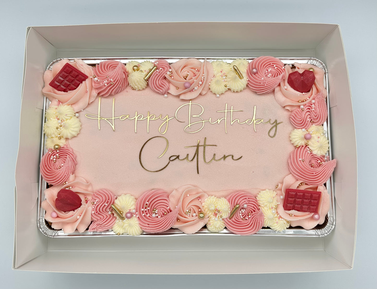 Pink birthday sheet cake with gold lettering, decorated with piped buttercream swirls, edible pearls, gold accents, and chocolate embellishments.